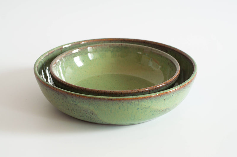 Two green-glazed RachaelPots Pasta Bowls, nestled together, showcasing a speckled finish and brown rims, ideal for serving pasta or noodles.