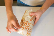 Bee's Wrap - Lunch 3 Pack - Honeycomb Print, featuring reusable sandwich and snack wraps with wooden bee button, held by a person wrapping a package.