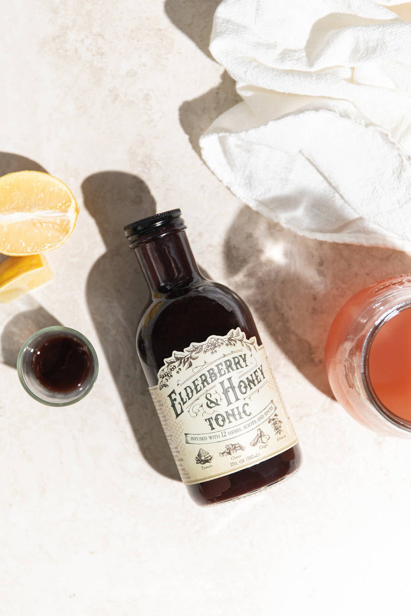 Roots And Leaves - Elderberry & Honey Tonic, 16 oz bottle with label, accompanied by a glass of brown liquid, on a surface.