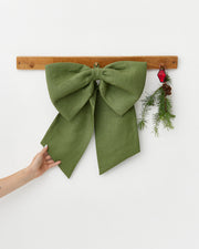 MagicLinen Forest Green Linen Decorative Bow on a wooden wall rack with evergreen sprig and red ornament, highlighting festive decor. A hand with heart tattoo touches the bow.