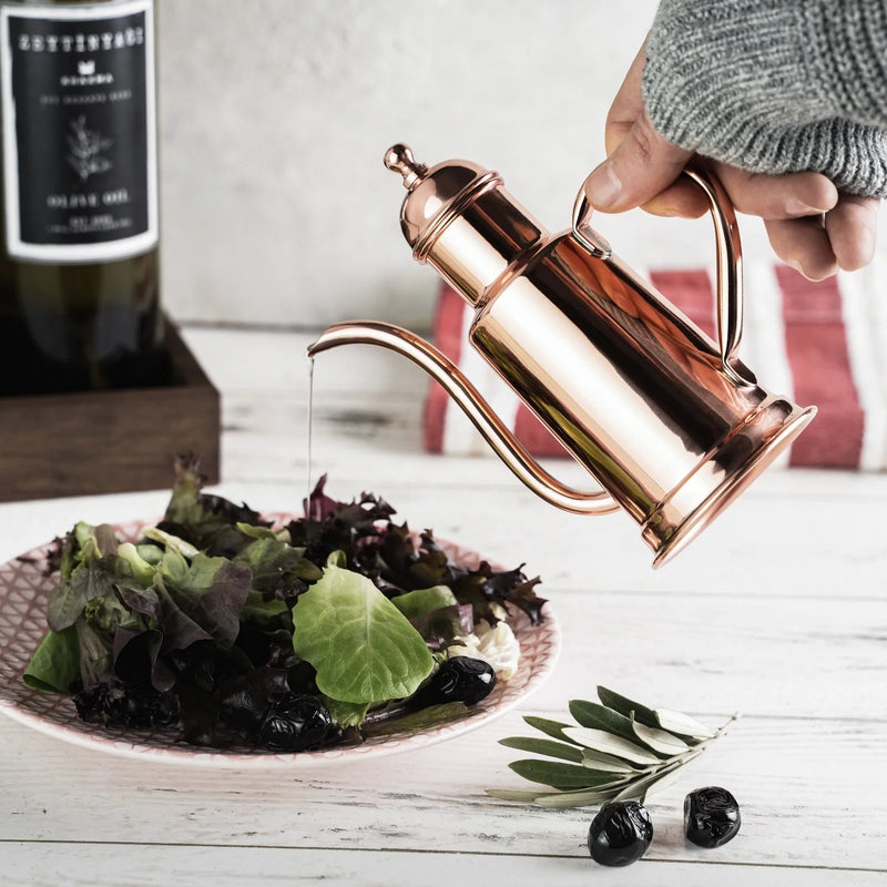 Hand holding ALFIQ Handmade Copper Olive Oil Dispenser, 600ml, drizzling oil onto salad greens, with olives and olive oil bottle in a rustic kitchen setting.