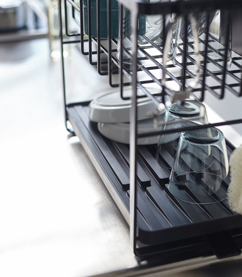 Two-Tier Dish Rack from Yamazaki Home features a black metal frame with a drip tray, drying glasses, plates, and cups on a stainless-steel countertop.