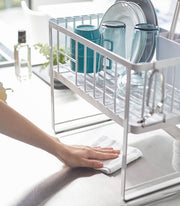 Yamazaki Home | Two-Tier Dish Rack, White, on a countertop with clean dishes; a hand wipes the surface beneath, showcasing its efficient design for kitchen organization.