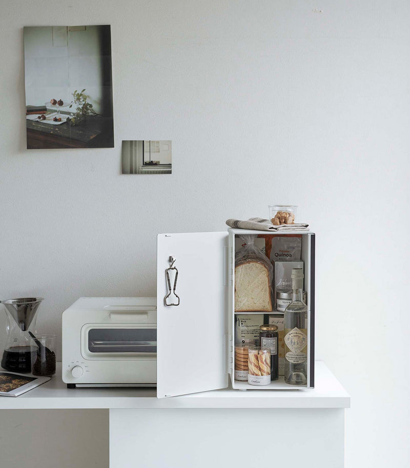 Yamazaki Home vertical steel bread box, black, keeps bread fresh on a minimalist kitchenette with coffee dripper, toaster-oven, and open mini-fridge displaying various foods.