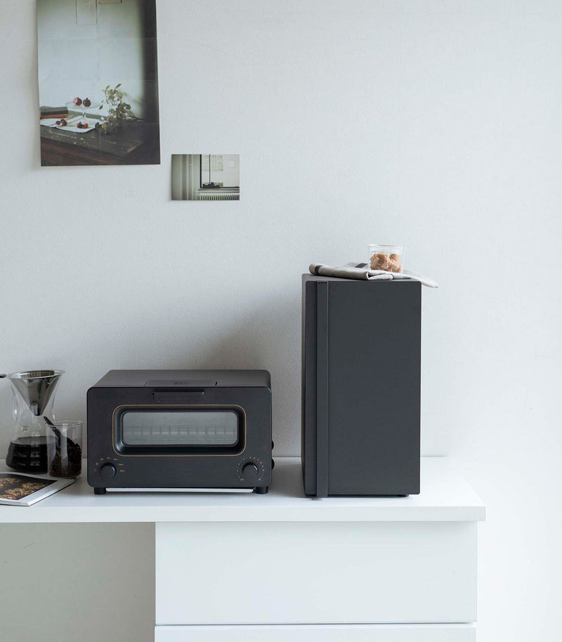 Yamazaki Home | Bread Box, Vertical, Steel, Black, sits amid a sleek kitchen setup, enhancing the decor and organizing space, perfect for keeping baked goods fresh.