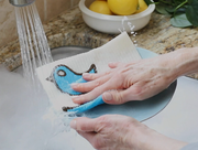 Three Bluebirds Swedish Dishcloths in action; a person washes a plate with a blue towel under running water.