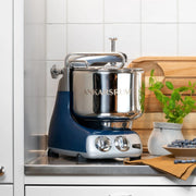 Ankarsrum stand mixer on a kitchen counter with a stainless-steel bowl, accompanied by a cutting board, potted mint, and blueberries.