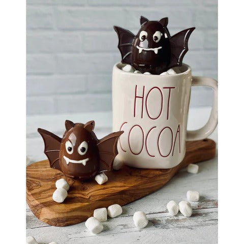 Chocolates Unlimited by Denise | Halloween Bat Hot Chocolate Bombs: Two chocolate spheres with bat wings, eyes, and fangs, surrounded by mini marshmallows near a HOT COCOA mug.