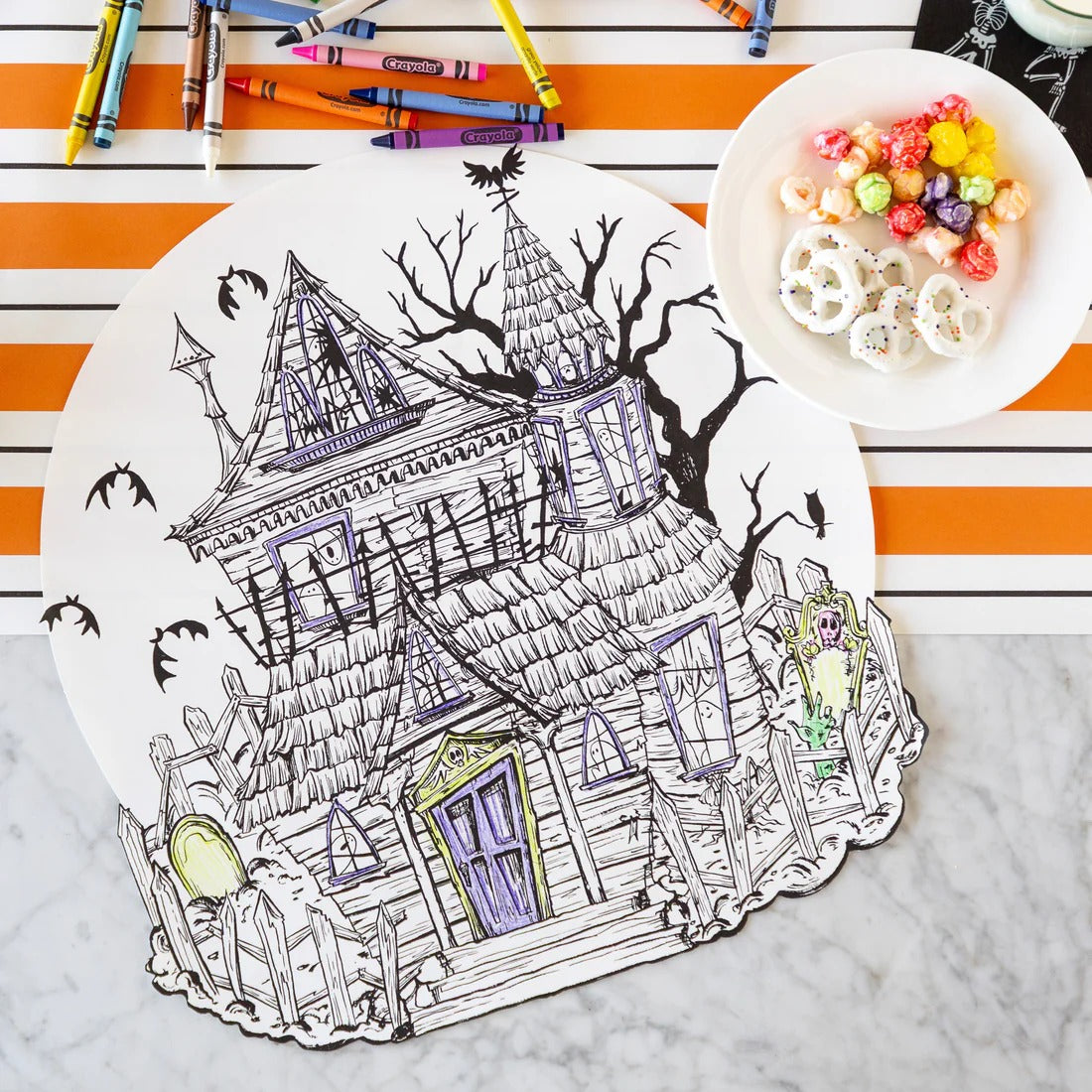 Hester & Cook | Haunted House Coloring Placemat on a countertop, partially colored with crayons, accompanied by a plate of candied popcorn and yogurt-covered pretzels.