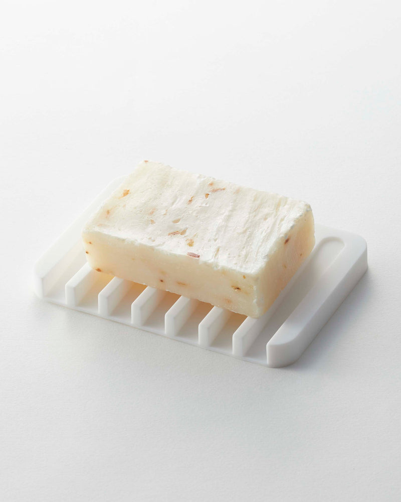 Yamazaki Home | Flow Self-Draining Soap Tray: Off-white bar soap with flecks on a black silicone tray, featuring raised ridges for effective drainage and drying.
