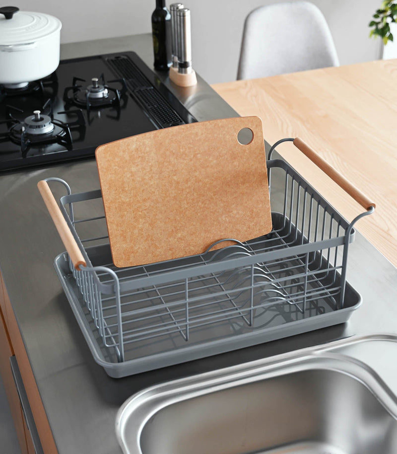 Yamazaki Home | Steel + Wood Dish Rack with Drainer Spout on a stainless-steel counter, holding a cutting board; features a stylish two-tiered design with wooden handles.