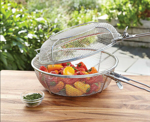 The 3 in 1 Grilling Basket by Fox Run Brands, packed with fresh and colorful vegetables including red and yellow peppers, is set on a wooden table outdoors. Beside the basket sits a small glass bowl filled with chopped herbs. The lush green plants in the background create an ideal setting for both indoor and outdoor cooking.