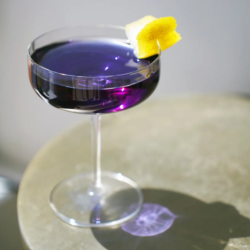 Bormioli Jazz Cocktail Coupe, 10.25 oz, filled with deep purple cocktail, garnished with lemon peel, sunlight casting a purple reflection on the table. Set of 4.