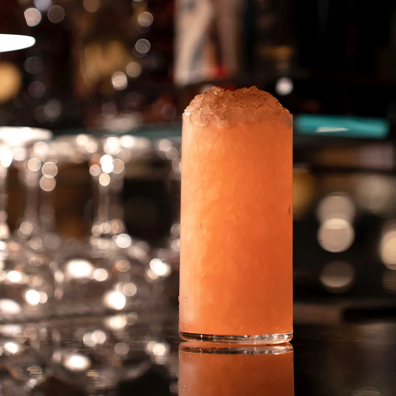 Bormioli highball glass filled with a bright orange slushy cocktail, set on a bar counter amidst soft bar light ambiance.