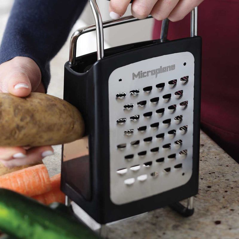Microplane | 4-Sided Professional Box Grater on counter; person grating potato, carrot and cucumber nearby, showcasing versatile, sharp blades ideal for diverse kitchen tasks.
