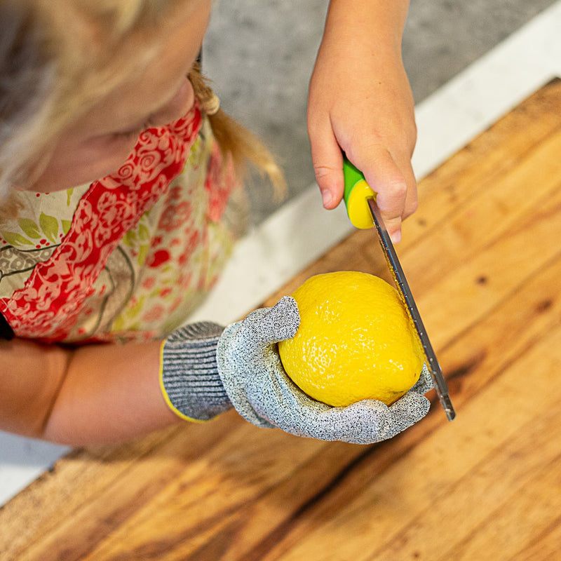 Child using Microplane | Cut Resistant Kitchen Safety Gloves for Kids, zesting a lemon with a handheld zester on a wooden board in a kitchen.