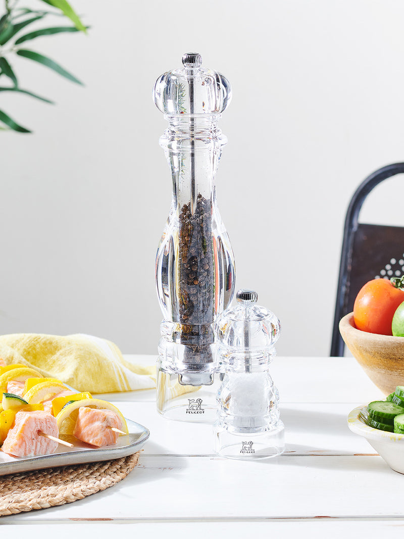 Peugeot | Paris Nancy Pepper Mill, Acrylic, 12 inches, on a white table with a salt grinder, salmon skewers, and fresh produce, showcasing culinary elegance.