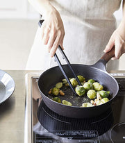 Person uses Yamazaki Home | Tower Floating Tweezer Tongs, Silicone, to sauté Brussels sprouts on induction cooktop, showcasing their drip-free floating design and practicality.