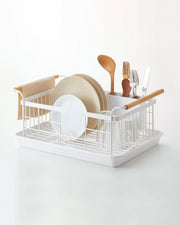 Yamazaki Home | Steel + Wood Dish Rack with Drainer Spout, featuring a sleek design with wooden handles, utensil caddy, and drip tray, holding assorted dishes and utensils.