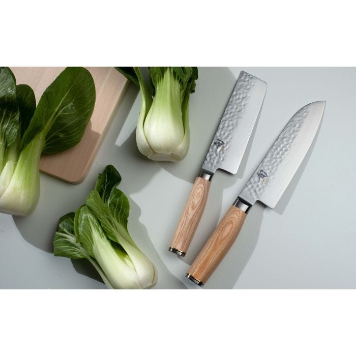 Shun Premier Blonde Santoku 7 knife with hammered finish and light handle, displayed on a wooden board with four baby bok choy and another Japanese knife.