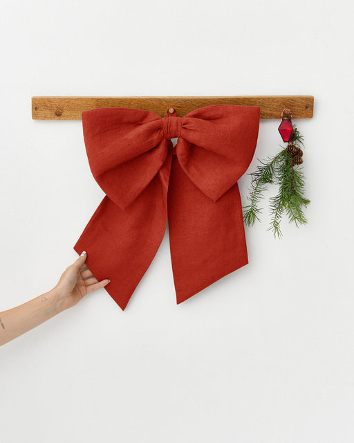 MagicLinen | Linen Decorative Bow, Red, elegantly draped on a wooden rail, with a hand lifting its tail, adding a festive touch to your space.