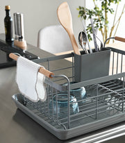Yamazaki Home | Steel + Wood Dish Rack with Drainer Spout on a kitchen counter, featuring utensils, bowls, and a hanging dish cloth, showcasing style and functionality.