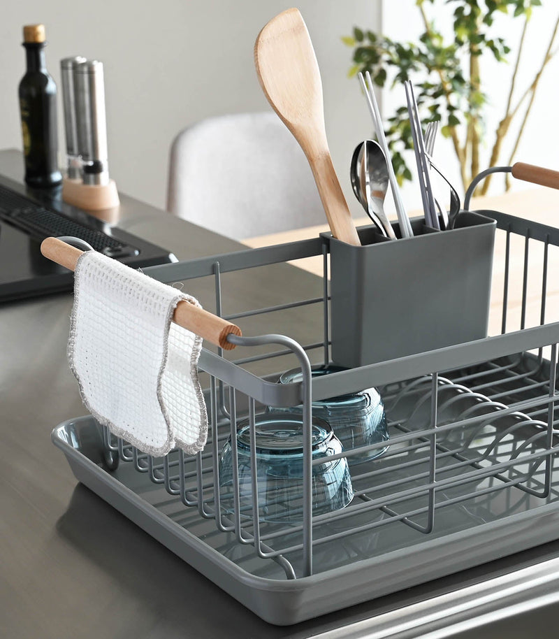 Yamazaki Home | Steel + Wood Dish Rack with Drainer Spout on a kitchen counter, featuring utensils, bowls, and a hanging dish cloth, showcasing style and functionality.