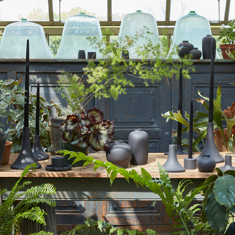 Rustic display with The Floral Society's Daily Plate, Sand, among potted plants, matte-black vessels, and candles, highlighting its versatility as a candle base or catchall.