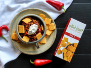 Southern Straws | Cheese Straws accompany a hearty chili bowl, showcasing the Spicy variety’s packaging, surrounded by fresh chili peppers and a rustic dining setup.