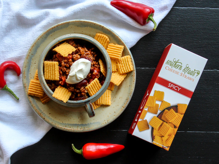 Southern Straws | Cheese Straws beside a chili con carne mug, topped with crackers, showcasing hand-crafted, spicy cheese wafers from a family recipe, ideal for snacking.