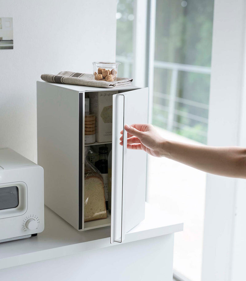 Yamazaki Home Bread Box showcased in a minimalist kitchen, emphasizing its vertical steel design for keeping baked goods fresh, with a fridge and toaster-oven nearby.
