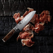 Shun | Premier Santoku Walnut, 7 knife with hammered finish, rests on cured ham slices atop a charred wooden surface, highlighting its elegance and craftsmanship.
