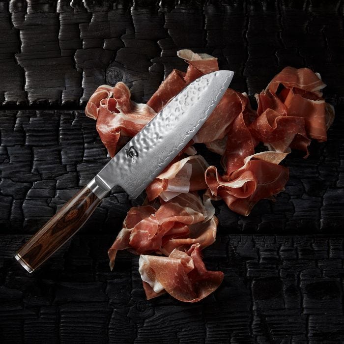Shun | Premier Santoku Walnut, 7 knife with hammered finish, rests on cured ham slices atop a charred wooden surface, highlighting its elegance and craftsmanship.