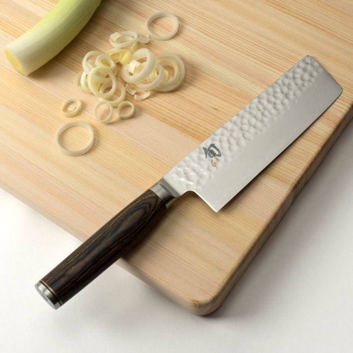 Shun | Premier Nakiri Walnut, 5.5 knife on a cutting board with sliced leek, showcasing its hammered finish and dark wood handle, ideal for precise vegetable cutting.