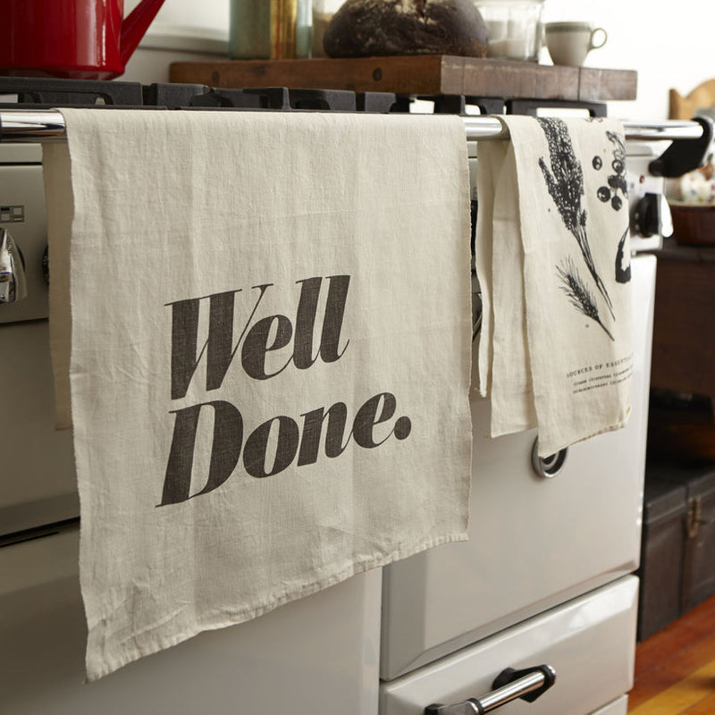 Sir|Madam Linen Tea Towels, Wine Varietals, draped on oven, paired with cozy kitchen scene featuring a red kettle and a loaf of bread.