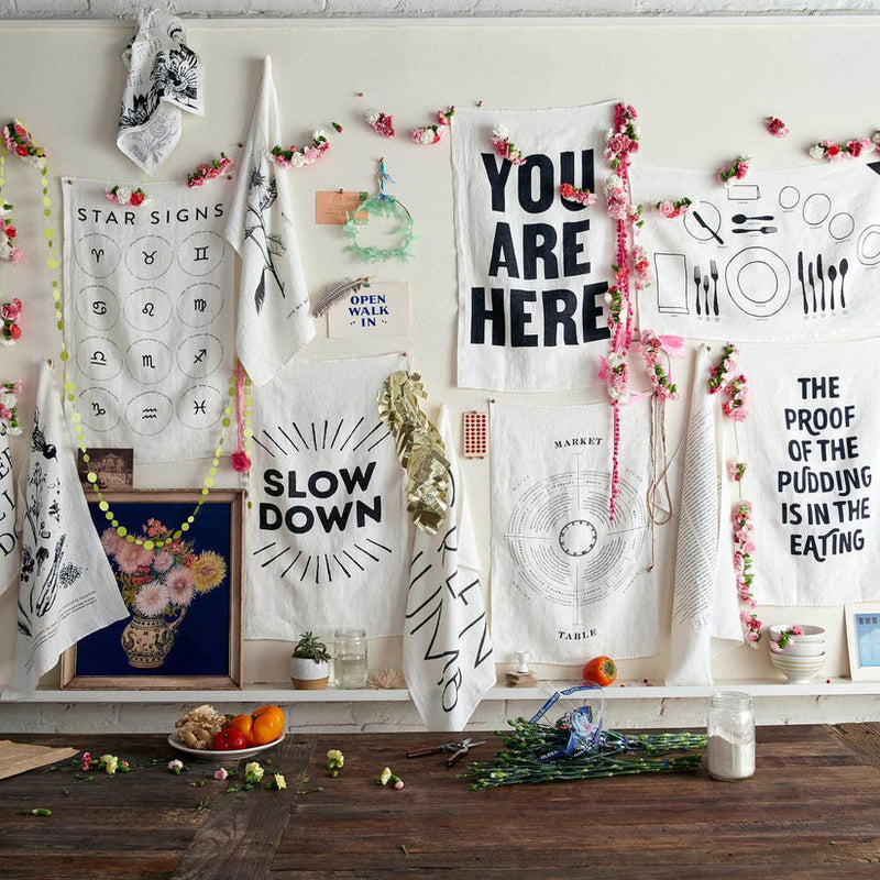Sir|Madam Linen Tea Towels, Slow Down hangs in a styled kitchen vignette, surrounded by printed fabrics, small art prints, and fresh flowers.