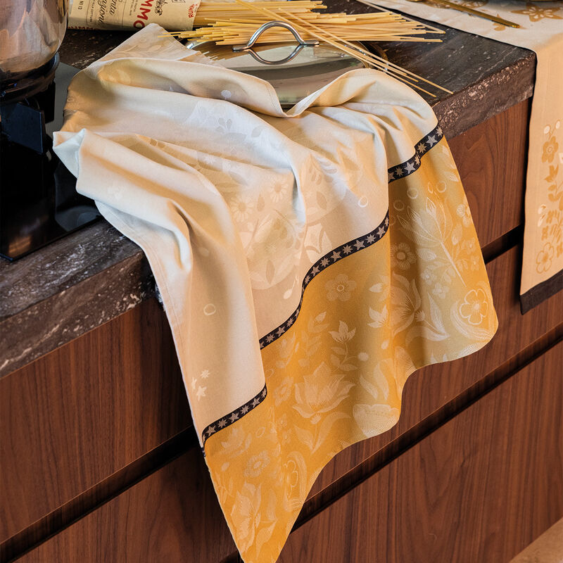 Le Jacquard Francais | Féerie Tea Towel, Yellow, draped over a kitchen counter, showcasing its floral pattern and festive charm beside pasta and cookware.