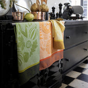 Le Jacquard Francais | Thanksgiving Tea Towel, Orange displayed on a black range cooker, enhancing a cozy kitchen setup with artichokes, cookware, and espresso machine nearby.