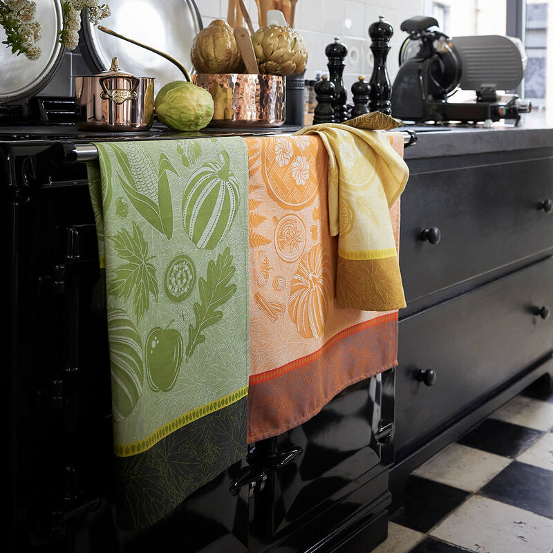 Le Jacquard Francais | Thanksgiving Tea Towel, Orange displayed on a black range cooker, enhancing a cozy kitchen setup with artichokes, cookware, and espresso machine nearby.