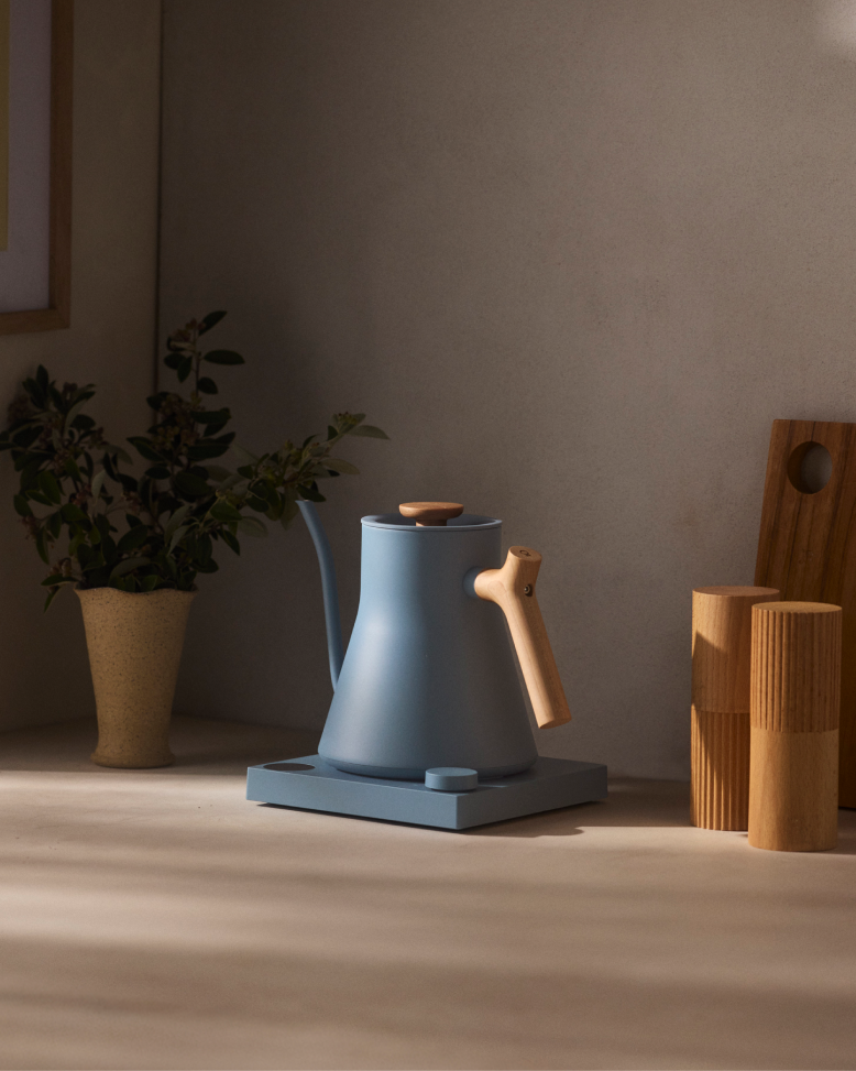 FELLOW | Stagg EKG Pro Electric Kettle in hazy blue, with maple accents, on a minimalist kitchen counter, ideal for precise coffee brewing.