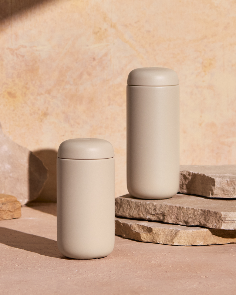 Two matte beige FELLOW | Carter Move Mugs, Sand Dune / 16 oz, with snug lids, designed for flavor preservation and spill-proof commuting, on stone slabs.