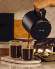 A hand pours coffee from the FELLOW | Aiden Precision Coffee Maker, Matte Black, into clear cups, showcasing precision brewing on a kitchen countertop.