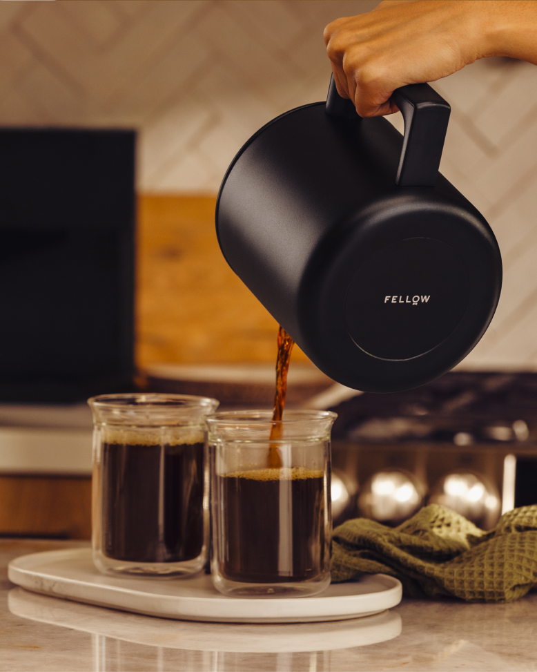 A hand pours coffee from the FELLOW | Aiden Precision Coffee Maker, Matte Black, into clear cups, showcasing precision brewing on a kitchen countertop.