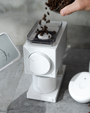 A hand pours beans into the FELLOW | Ode Brew Grinder Gen 2, Matte White, with a visible grind-size dial, set on a stone countertop alongside a scale and timer.