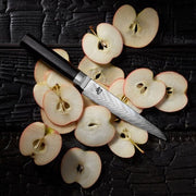 Shun | Dual Core Utility & Butchery Knife, 6 inches, displayed on charred wood with apple slices, features a Damascus-style blade and dark handle.