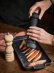 Hands using the Peugeot | Paris Nature Collection Pepper Mill, Nature Black, to season sweet potato wedges on a tray, highlighting sustainable design and adjustable grind setting.