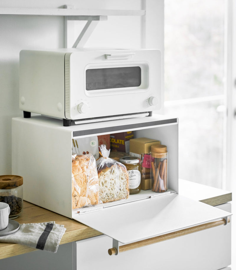 White two-tier Yamazaki Home Simple Steel Bread Box with toaster oven on top, storing bread, jam, and cocoa inside; coffee jar and cup nearby on a wooden counter.