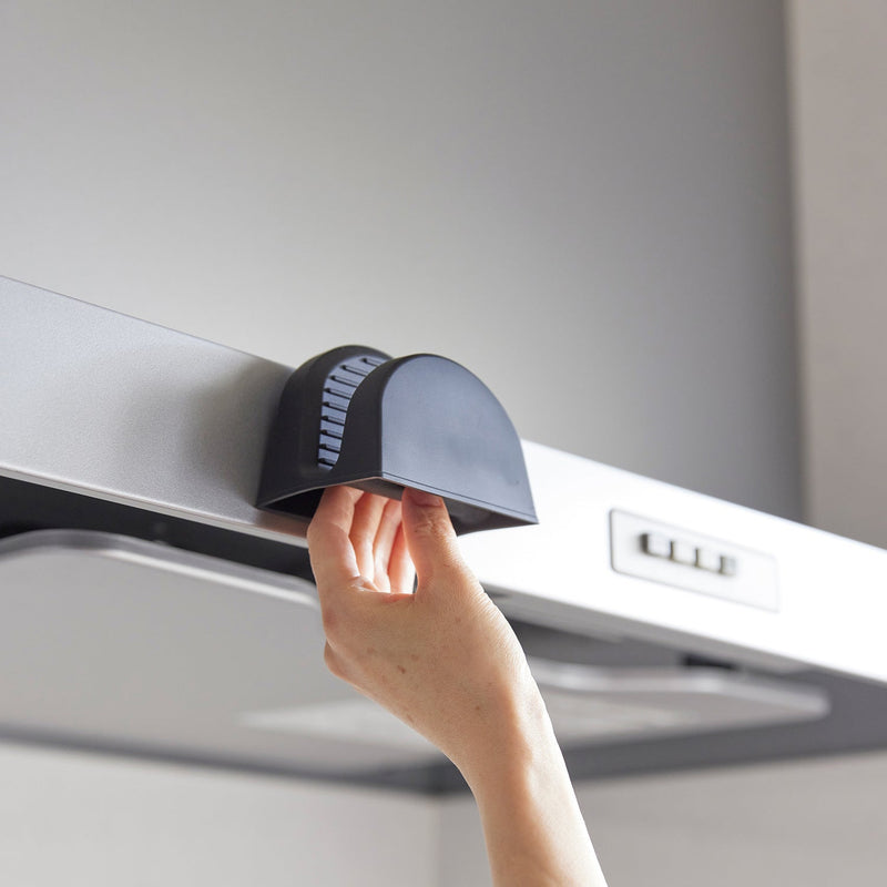 Hand pulling down a black magnetic pot holder from a kitchen range hood, showcasing the Yamazaki Home Silicone Pot Holders, ideal for safe handling in the kitchen.