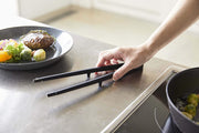 Yamazaki Home | Tower Floating Tweezer Tongs, Silicone, resting on a stainless-steel counter next to an induction cooktop with Brussels sprouts in a frying pan.