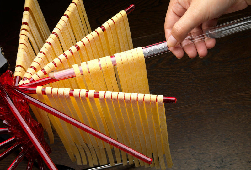 Fresh pasta ribbons hang to dry on the Marcato | Tacapasta Pasta Drying Rack, Red, with a hand adjusting a spoke for optimal drying.